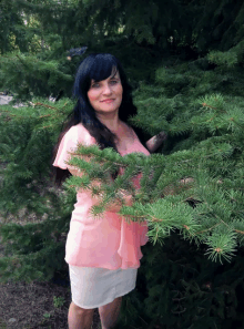 a woman in a pink top and white skirt is standing in front of a pine tree