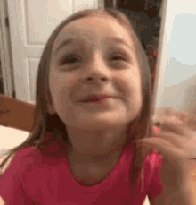 a little girl in a pink shirt is smiling and holding a cell phone in her hand .
