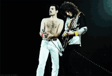a man without a shirt is singing into a microphone and another man is playing a guitar .
