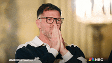 a man wearing glasses is covering his mouth with his hands and the nbc logo is visible in the corner