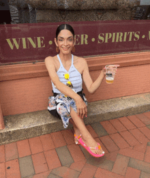 a woman sits on a brick sidewalk holding a glass of beer in front of a wine and beer store