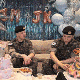 two men in military uniforms are sitting on a couch in front of balloons and a birthday cake .