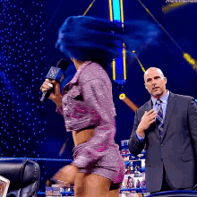 a woman in a purple outfit is holding a microphone while a man in a suit looks on