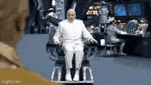 a bald man in a white suit is sitting in a chair in a room .