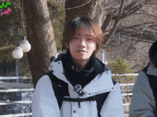 a person wearing a white jacket and a black scarf stands in front of a tree