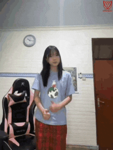 a girl is standing in front of a pink chair and a clock on the wall