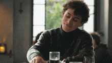 a man with curly hair sits at a table with a glass of beer