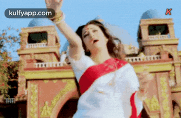 a woman in a white and red saree is dancing in front of a building with her arms in the air .