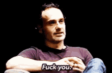 a man in a purple shirt is sitting in a chair and says " fuck you "