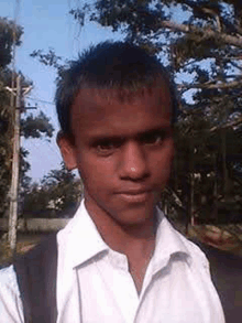 a young man in a white shirt is standing in front of a tree and looking at the camera .
