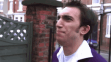 a man wearing a purple sweater and white shirt is standing in front of a brick wall