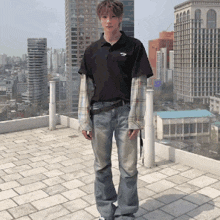 a young man wearing a black polo shirt and jeans stands on a rooftop