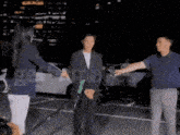 a man in a suit holds a woman 's hand in a parking lot while another man holds a microphone