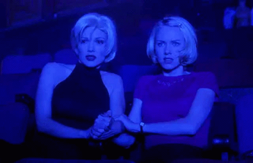 two blonde women hold onto each other in a theatre watching the stage. they are bathed in blue light