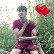 a man in a red shirt and shorts is sitting in the grass with two red hearts behind him