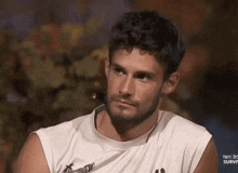 a man with a beard is wearing a white sleeveless shirt and looking at the camera .
