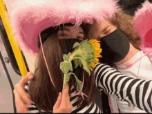 a woman wearing a pink hat and a black mask holds a sunflower in front of her face