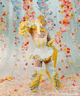 a drag queen is standing in front of a wall of flowers wearing a white and yellow dress and gloves .
