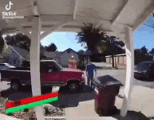 a red truck is parked under a canopy in a driveway next to a trash can .