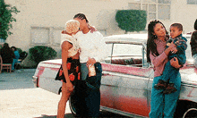a woman holding a child stands next to a man and woman kissing in front of a car