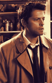 a man in a trench coat and tie is standing in front of a shelf with bottles on it