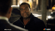 a man in a black jacket is talking to another man in a suit with the nbc logo behind him