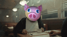 a man wearing a pig mask sits at a table with a cup of coffee