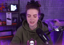 a woman wearing a green hoodie with a skull on it stands in front of a shure microphone