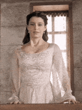 a woman in a white dress with pearls on the sleeves is standing in front of a window