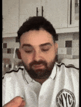 a man with a beard wearing a white nyc polo shirt is sitting in a kitchen .