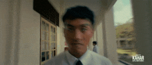 a man in a white shirt and black tie is walking down a hallway with kahar written on the bottom right