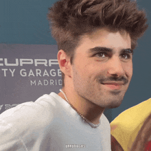 a man wearing a pearl necklace smiles in front of a cupra ty garage madrid sign
