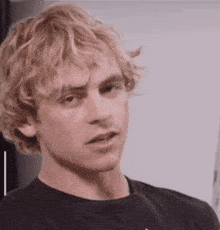 a young man with blonde curly hair is wearing a black shirt and making a funny face .