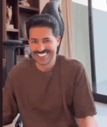 a man with a mustache is smiling and wearing a brown shirt .