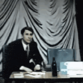 a man in a suit and tie is sitting at a table with a bottle of wine on it