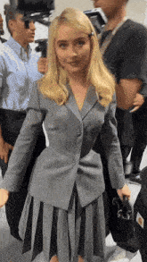a woman in a gray suit and pleated skirt stands in front of a camera