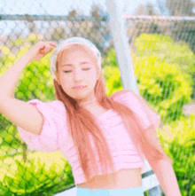 a woman wearing a pink crop top and a headband stands in front of a chain link fence