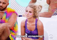 a woman in a purple top is screaming while sitting next to a man in a rainbow outfit .
