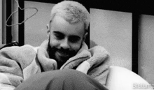 a black and white photo of a man with a beard sitting on a bed with a blanket .
