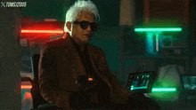 a man in a suit and sunglasses sits in front of a neon sign that says xtoms / 2005