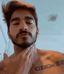 a shirtless man with a beard and a tattoo on his chest is shaving his beard .