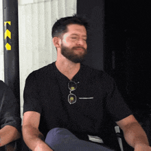 a man with a beard wearing a black t-shirt and sunglasses sitting in a chair .