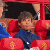 a man in a blue jacket is sitting in red seats with the manchester united logo on them