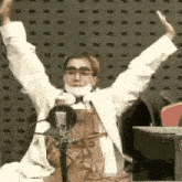 a man wearing a mask and glasses is sitting in front of a microphone with his arms in the air .
