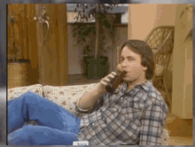 a man in a plaid shirt is drinking a beer while sitting on a couch