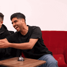 two men are sitting on a red couch laughing with a bell on a table
