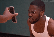 a man in a white tank top is looking at someone holding a cell phone in front of his face