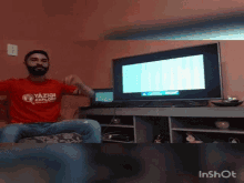 a man wearing a red shirt that says yazigi explorer is sitting in front of a television
