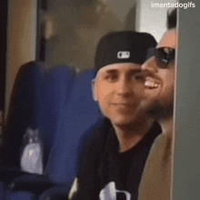 a man wearing a hat and sunglasses applauds while another man looks on