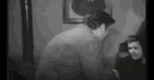 a black and white photo of a man standing next to a woman sitting on a couch .